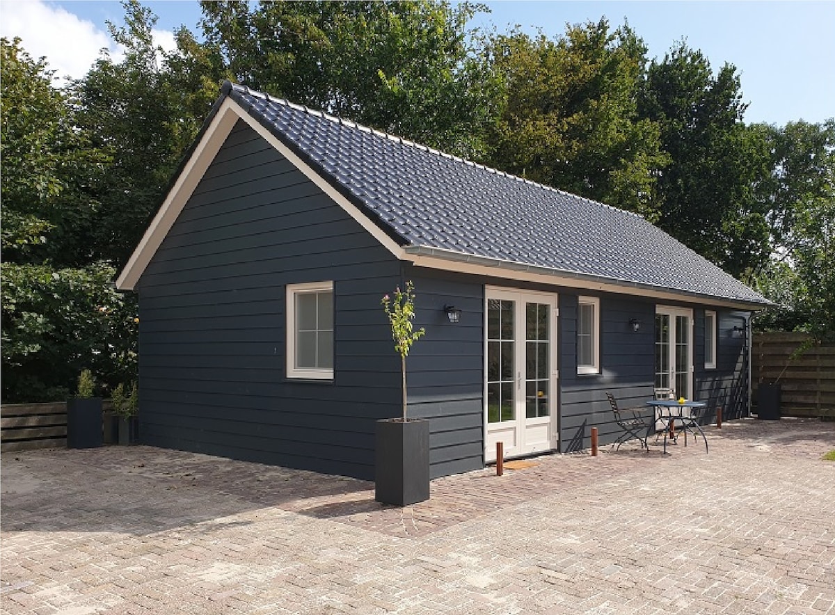 The exterior of the accommodation is shown, featuring a modern, dark blue wooden facade with a sloped roof. The facade includes large glass doors that open to a paved terrace area, where a small outdoor table and chairs are situated. Lush greenery surrounds the building.