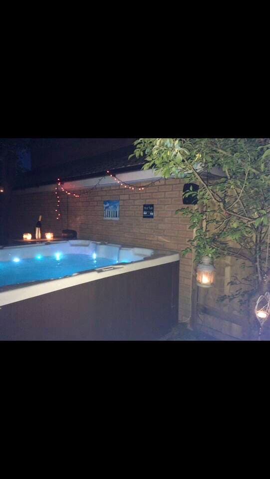 A private hot tub is surrounded by a low wooden wall, featuring illuminated blue lights. Soft, ambient lighting is provided by lanterns and string lights above. Vegetation adds a natural element, creating a tranquil outdoor space for relaxation in the evening.
