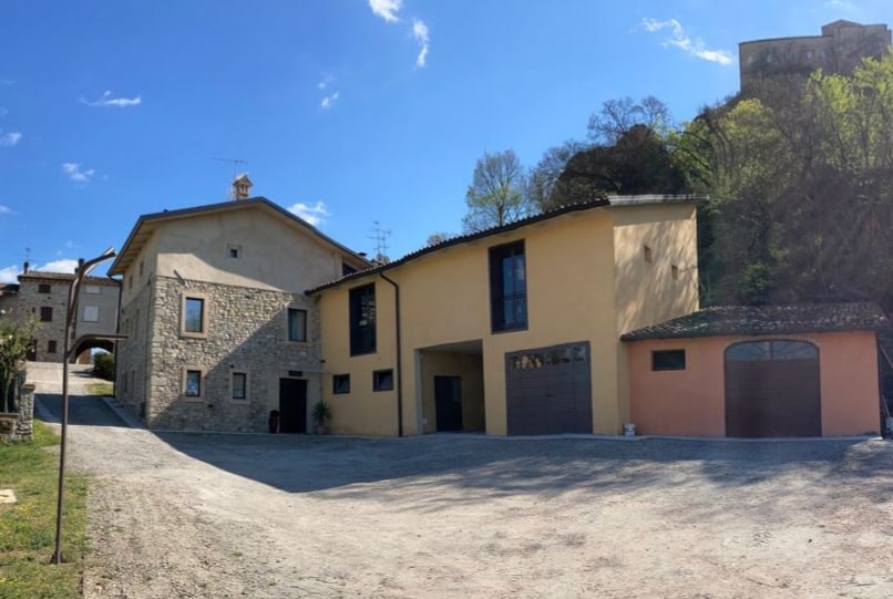 A scenic view of a rustic country house surrounded by greenery and hills. The building's exterior features a combination of stone and rustic painted surfaces. Parking spaces are visible in front, along with a gravel driveway leading to the entrance.