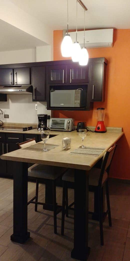 A modern kitchen space features dark cabinetry and a light-colored countertop. Three pendant lights illuminate a small dining area with two high stools. Kitchen appliances, including a microwave and a blender, are neatly arranged on the counter. An orange accent wall adds a vibrant touch.