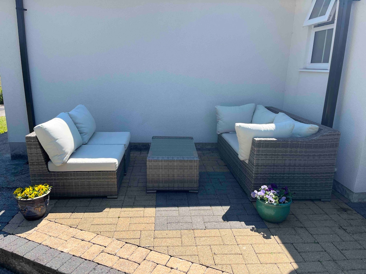An outdoor seating area features two comfortable, cushioned sofas arranged around a glass coffee table. Potted flowers in colorful pots add a touch of nature, complementing the stone-paved patio, which is illuminated by natural light.