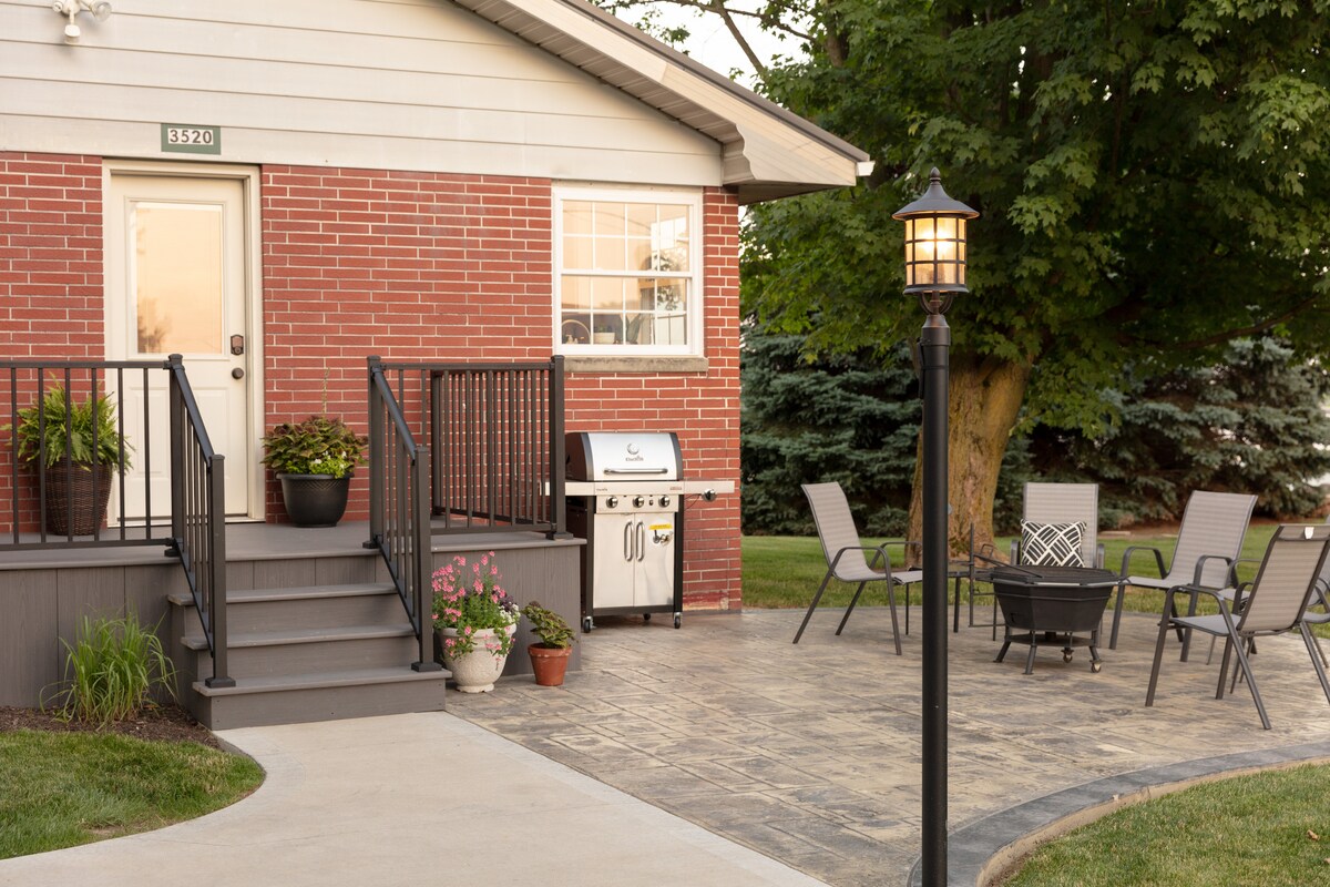 The exterior features a modern ranch house with a welcoming brick facade. A patio area is equipped with a round table and six chairs, while a grill sits nearby. A lamp post adds illumination, enhancing the outdoor space surrounded by greenery.