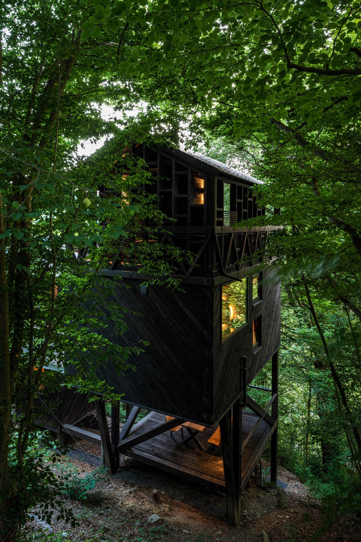 A modern treehouse structure is elevated on stilts, surrounded by lush green foliage. Large windows reveal warm light from within, while a wooden balcony offers a glimpse of outdoor space below. The design integrates with the natural environment, creating a serene and inviting presence.