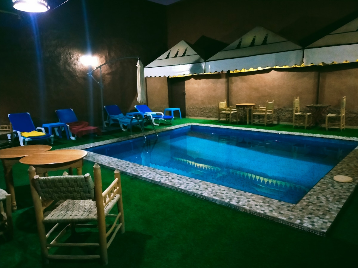 A serene swimming pool is framed by a landscaped area featuring green grass. Sun loungers and small tables are positioned around the pool, providing a relaxing space for guests. The warm lighting creates a calm ambiance, ideal for relaxation.