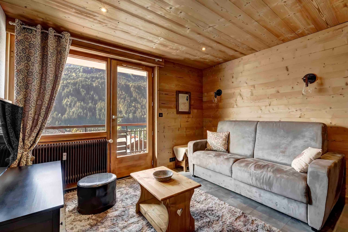 A warm and inviting living space is featured, with wooden paneling and a large window offering views of the surrounding mountains. A comfortable gray sofa sits adjacent to a small coffee table, and a dark rug adds texture to the floor.
