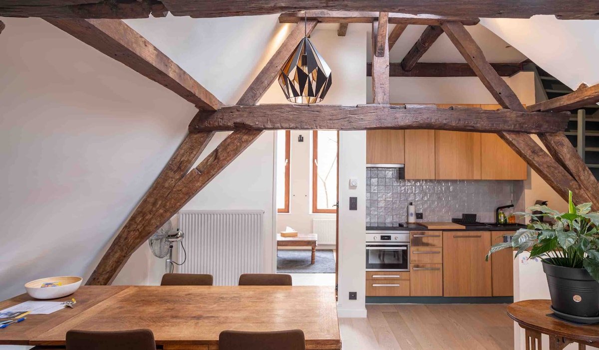Exposed wooden beams characterize the ceiling of the open living space, enhancing a modern kitchen with wood cabinetry and stainless steel appliances. A wooden dining table with upholstered chairs is positioned in the foreground, while a potted plant adds a touch of greenery.