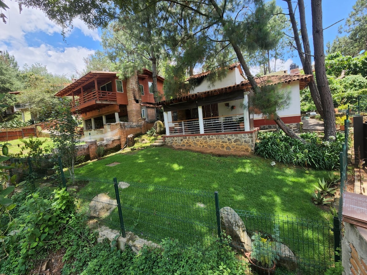 A charming cabin is nestled among trees, featuring rustic architecture with a stone foundation. The expansive yard is covered in lush green grass, bordered by a low fence and adorned with natural rocks. Nearby, two structures offer terraces overlooking the serene outdoor space.