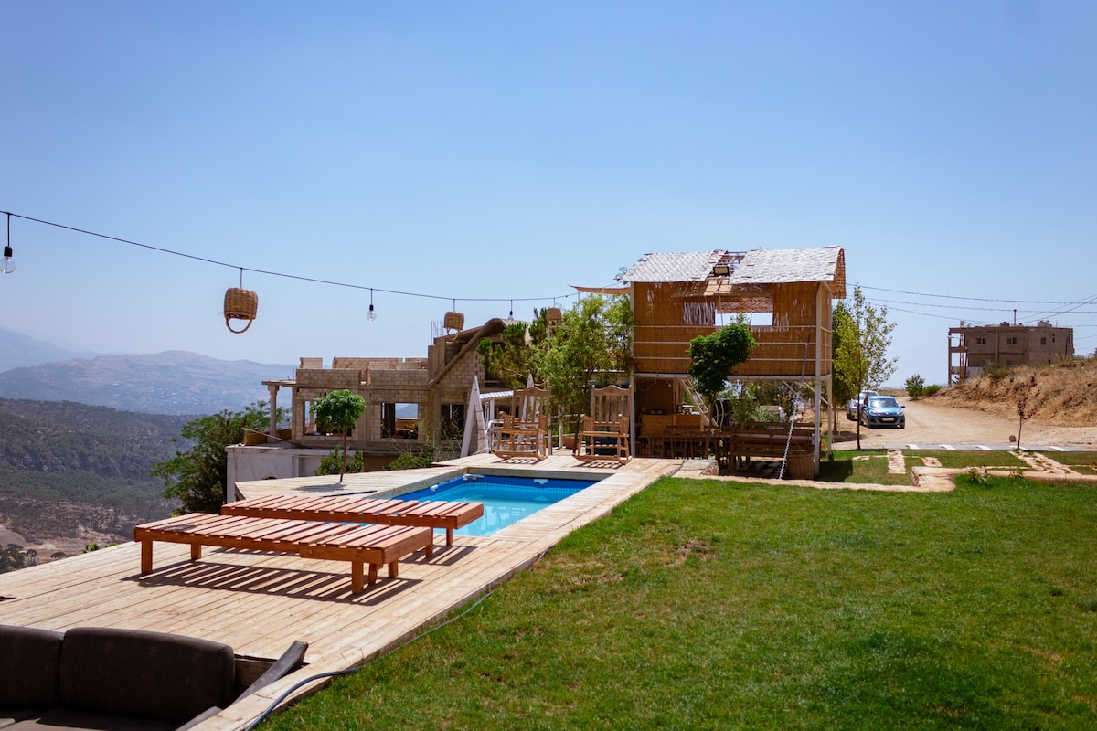 An open area features a wooden deck with lounge seating and a small pool, surrounded by grass. A treehouse adds charm, while a parking space is visible in the background. Scenic views of the Ain Dara village and distant hills are captured under a clear sky.