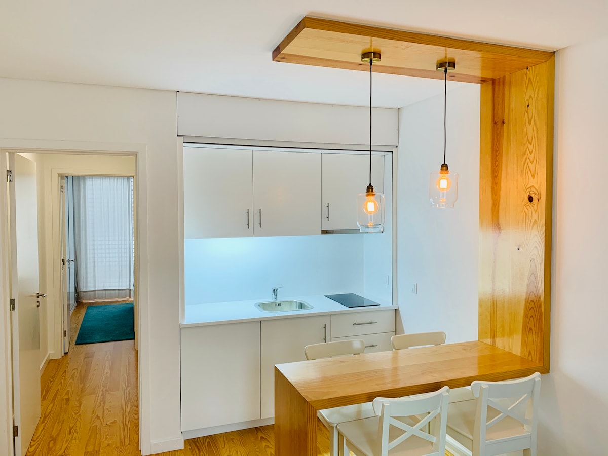 A modern kitchen is featured, showcasing sleek white cabinetry and a wooden accent above the dining area. A small sink and stovetop are visible, complemented by a light wood dining table with white chairs. Soft illumination is provided by two pendant lights.