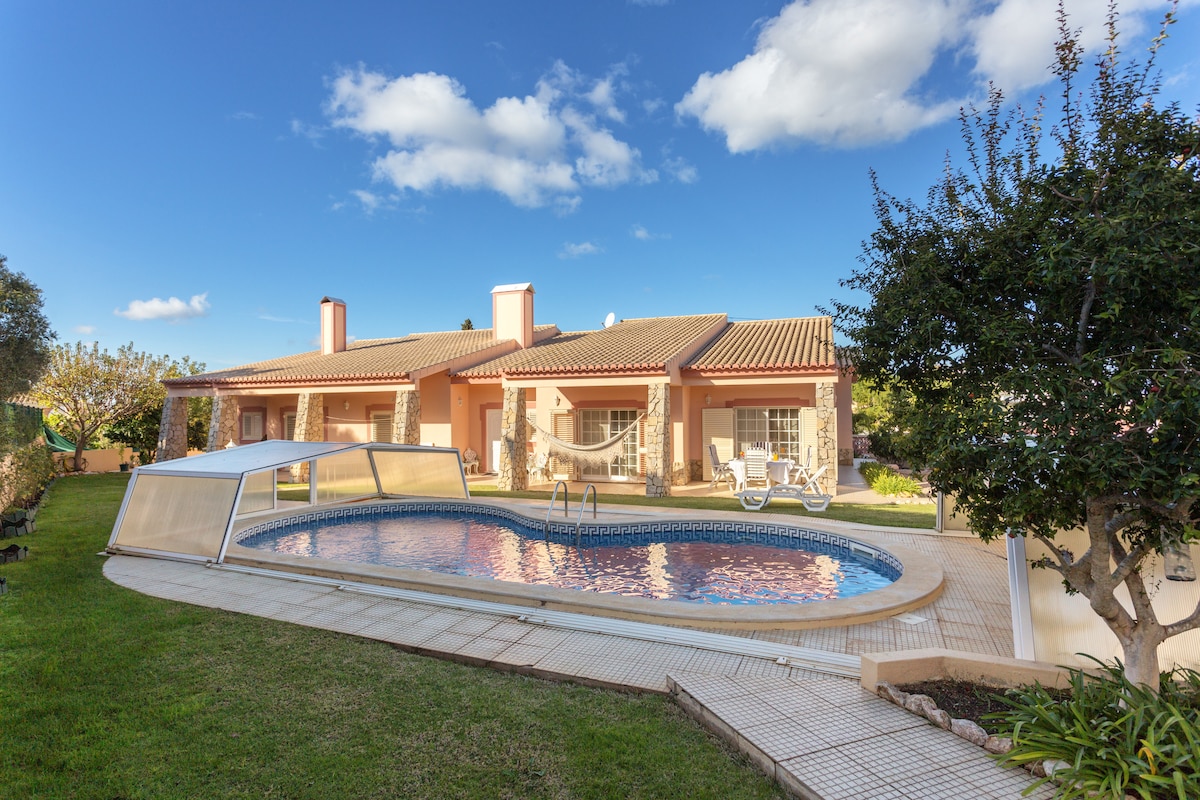 The exterior shows a spacious private pool surrounded by a well-maintained garden and patio area. The house features a warm design with multiple columns and a tiled roof. Lush greenery complements the serene setting under a clear blue sky with scattered clouds.