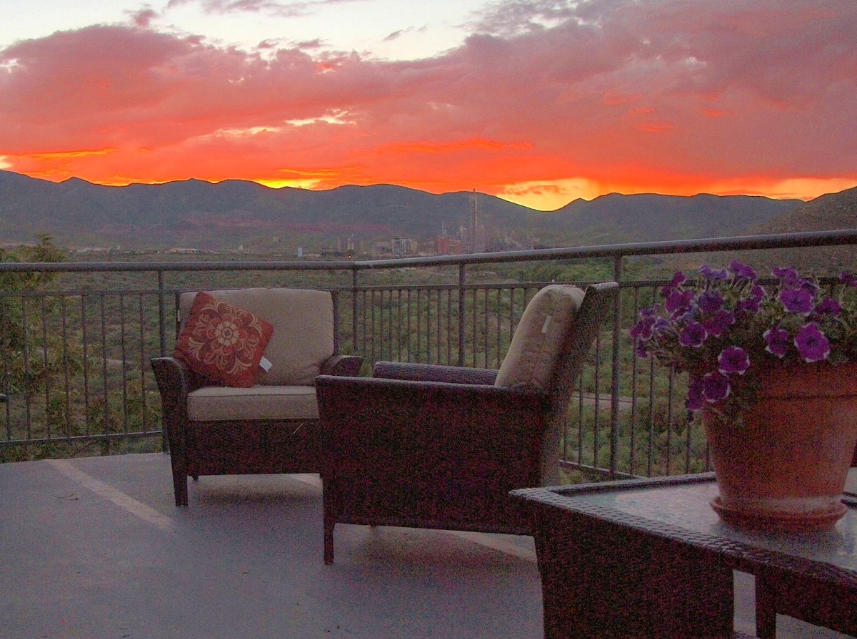 A tranquil outdoor view captures a vibrant sunset over rolling hills. A cozy seating arrangement with plush cushions is positioned on a patio, complemented by a potted plant filled with colorful flowers. The scene invites relaxation and appreciation of the natural beauty.