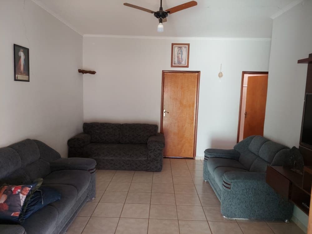 A living area is shown with two plush sofas facing each other, placed on tiled flooring. A ceiling fan is visible, providing ventilation. A wooden door leads to another room, while a wall-mounted frame adds a decorative touch.