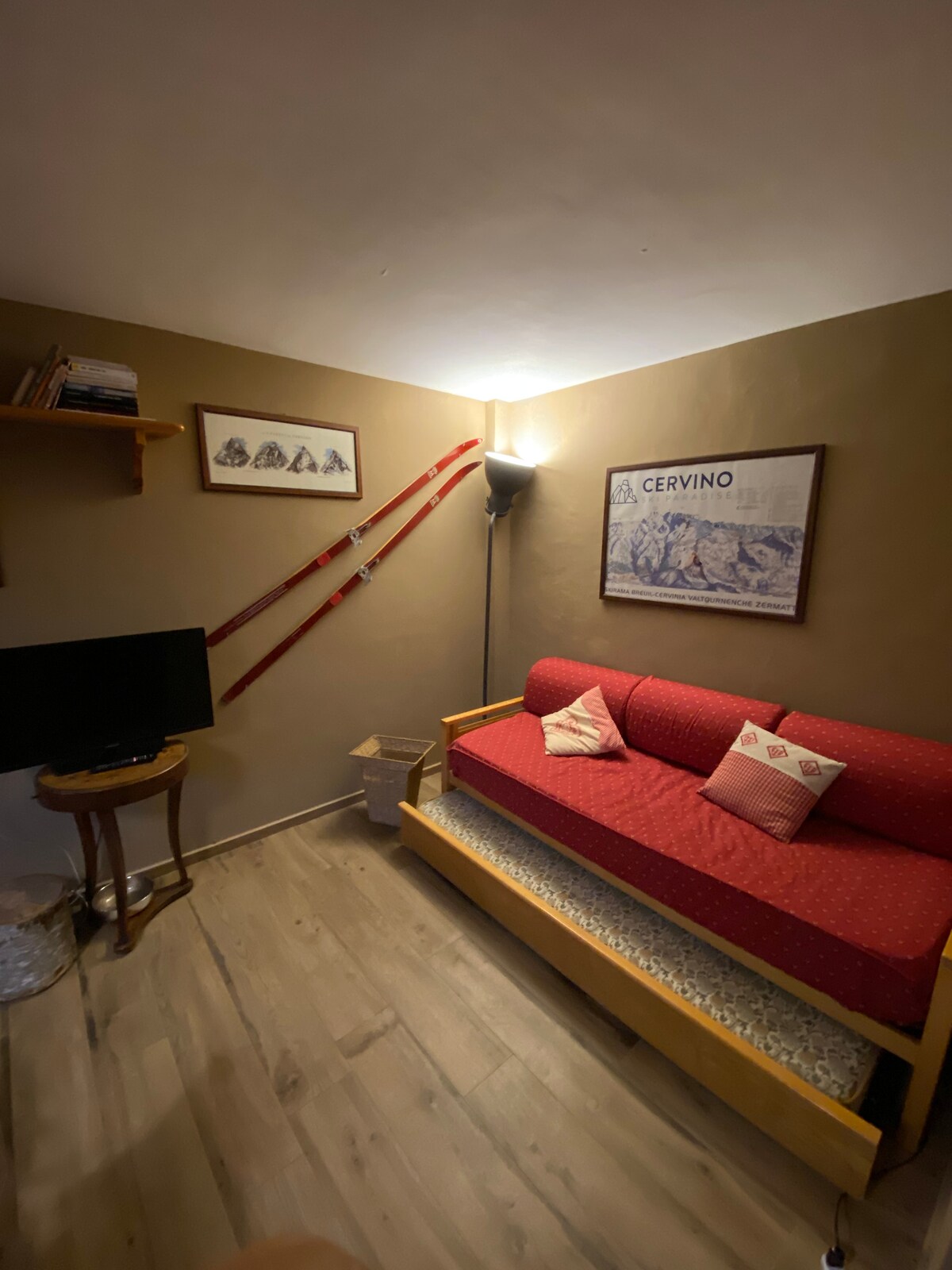 A cozy seating area features a red sofa bed adorned with patterned cushions. A TV stands on a small table nearby, and wall-mounted shelves display books. Vintage skiing equipment leans against the wall, enhancing the room's ambiance.
