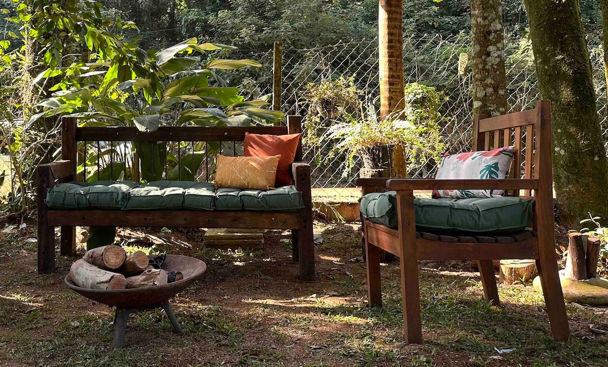 An outdoor seating area is surrounded by lush greenery, featuring two wooden couches adorned with green cushions and decorative pillows. A fire pit made of metal holds logs, offering a cozy gathering spot amidst nature. Sunlight filters through the trees, enhancing the serene atmosphere.