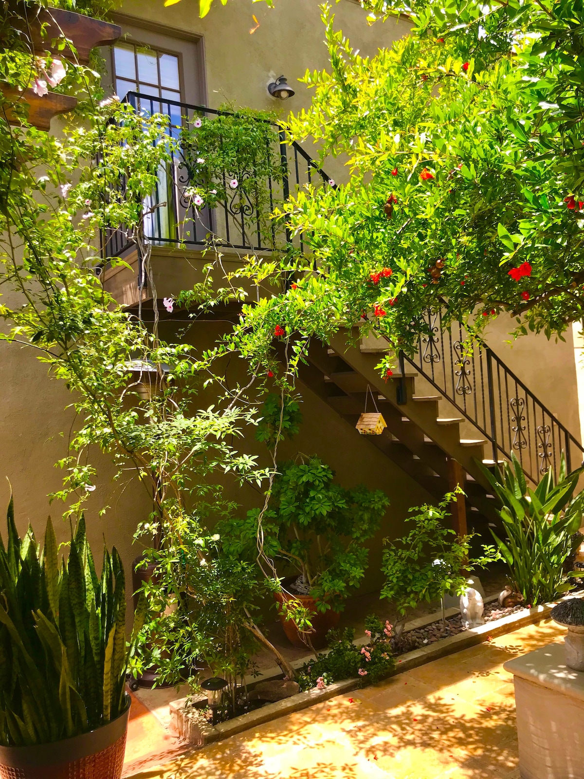 A charming outdoor staircase is framed by vibrant greenery and blooming flowers. Lush plants are positioned along the pathway, contributing to the serene atmosphere. The textured wall adds warmth to the scene, creating an inviting entrance to the upper level.
