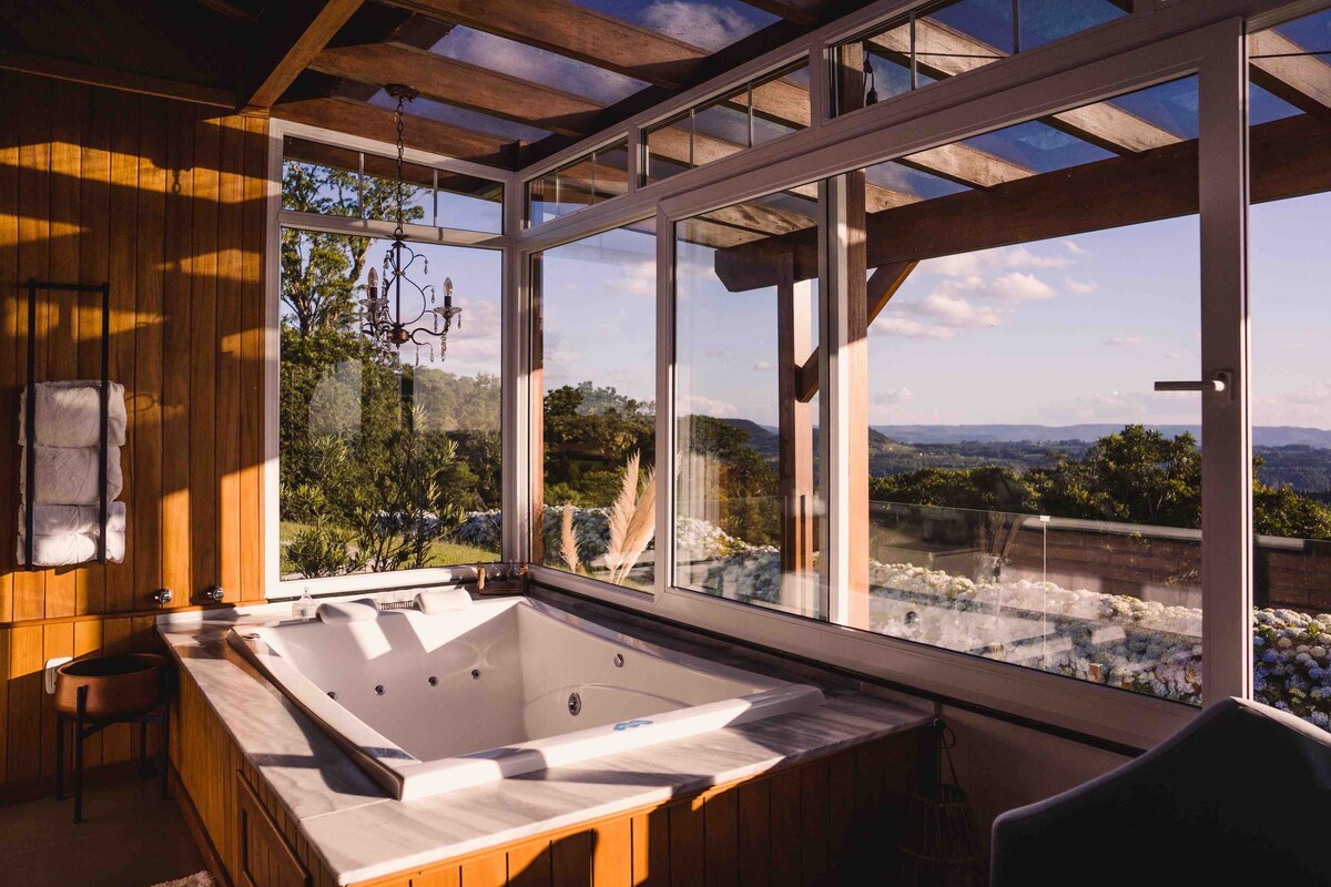 A spacious bathroom features a modern hot tub surrounded by large glass windows that provide stunning views of the surrounding landscape. Natural light illuminates the wooden accents and elegant chandelier, enhancing the tranquil atmosphere.