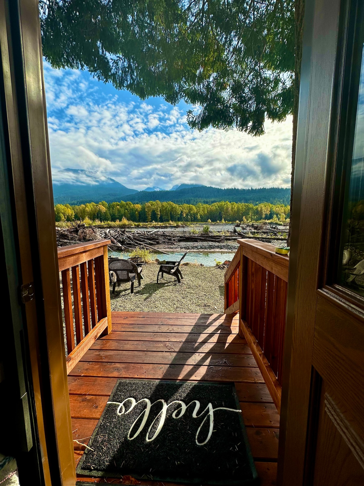 A view from the entrance features wooden steps leading to a riverfront area, with lounge chairs positioned on the gravel. Lush trees and distant mountains create a scenic backdrop, while a welcome mat greets guests at the threshold, inviting them into the tranquil environment.