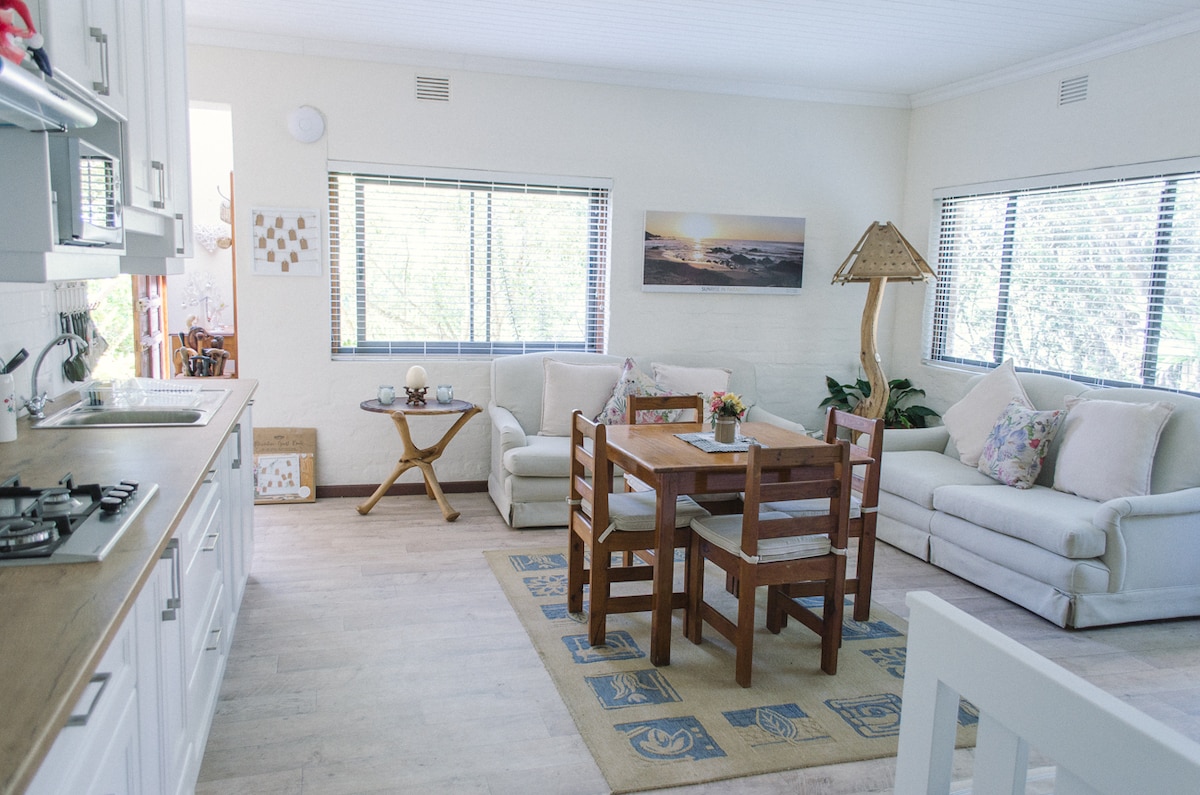 An open-plan living area features a wooden dining table surrounded by four chairs. A light-colored sofa is set against the wall, with a floor rug positioned beneath it. Large windows allow natural light to fill the space, enhancing the relaxed atmosphere.