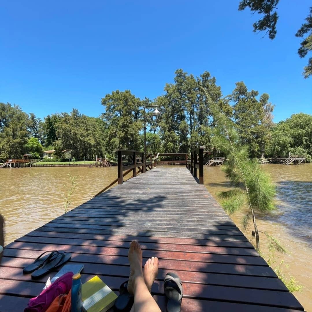 A wooden dock extends over the river, surrounded by lush greenery and tall trees. The calm water reflects the blue sky, creating a serene atmosphere. Soft shadows are cast on the dock, where a pair of sandals and a book can be seen resting.