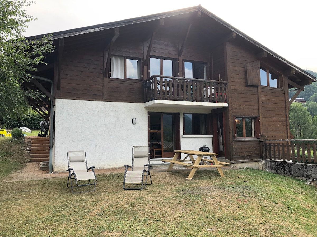 A wooden chalet is set against a natural backdrop, featuring a spacious patio with a picnic table and two lounge chairs. Large windows allow light to fill the interior, while a sloping roof and balcony add character to the structure.