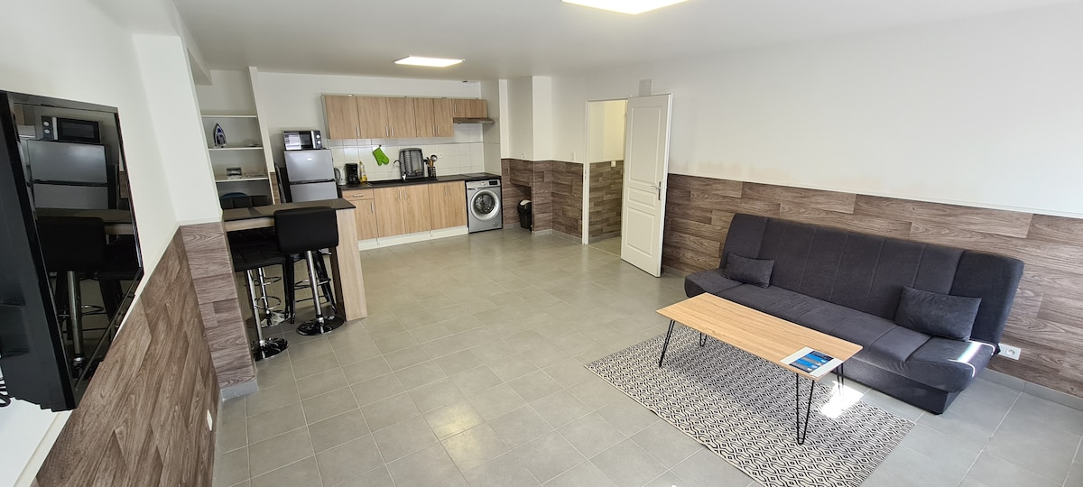 A spacious living area showcases a modern kitchen with wood cabinetry and essential appliances, including a washing machine. A dark couch is positioned along one wall, complemented by a coffee table on a textured rug. Natural light enters through a ceiling fixture and open space.