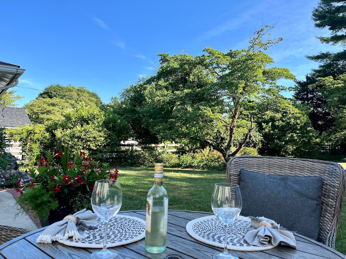 A dining table is set outdoors with two glasses and a bottle of wine. Lush greenery, including trees and flowering plants, serves as a backdrop under a clear blue sky, creating a serene environment for outdoor dining.