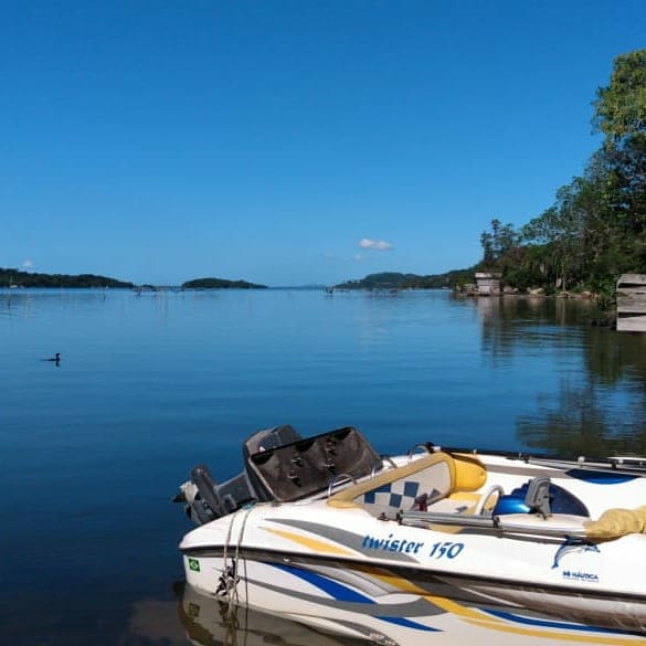 A small boat is moored at the edge of a serene body of water, reflecting clear blue skies. Gentle ripples disturb the surface, with distant islands visible on the horizon. Lush greenery borders the shoreline, enhancing the tranquil setting.