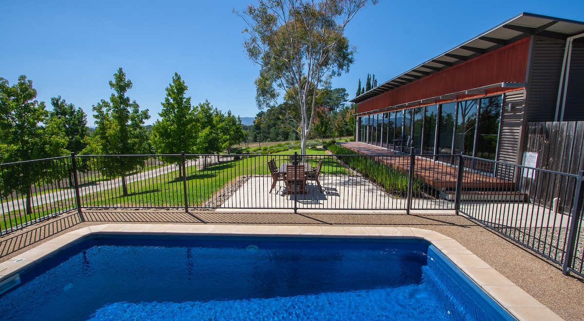 A private solar-heated swimming pool is surrounded by a secure fence, offering a clear view of the spacious outdoor area. Lush green trees line the property, with a patio and outdoor dining set visible adjacent to the guesthouse.