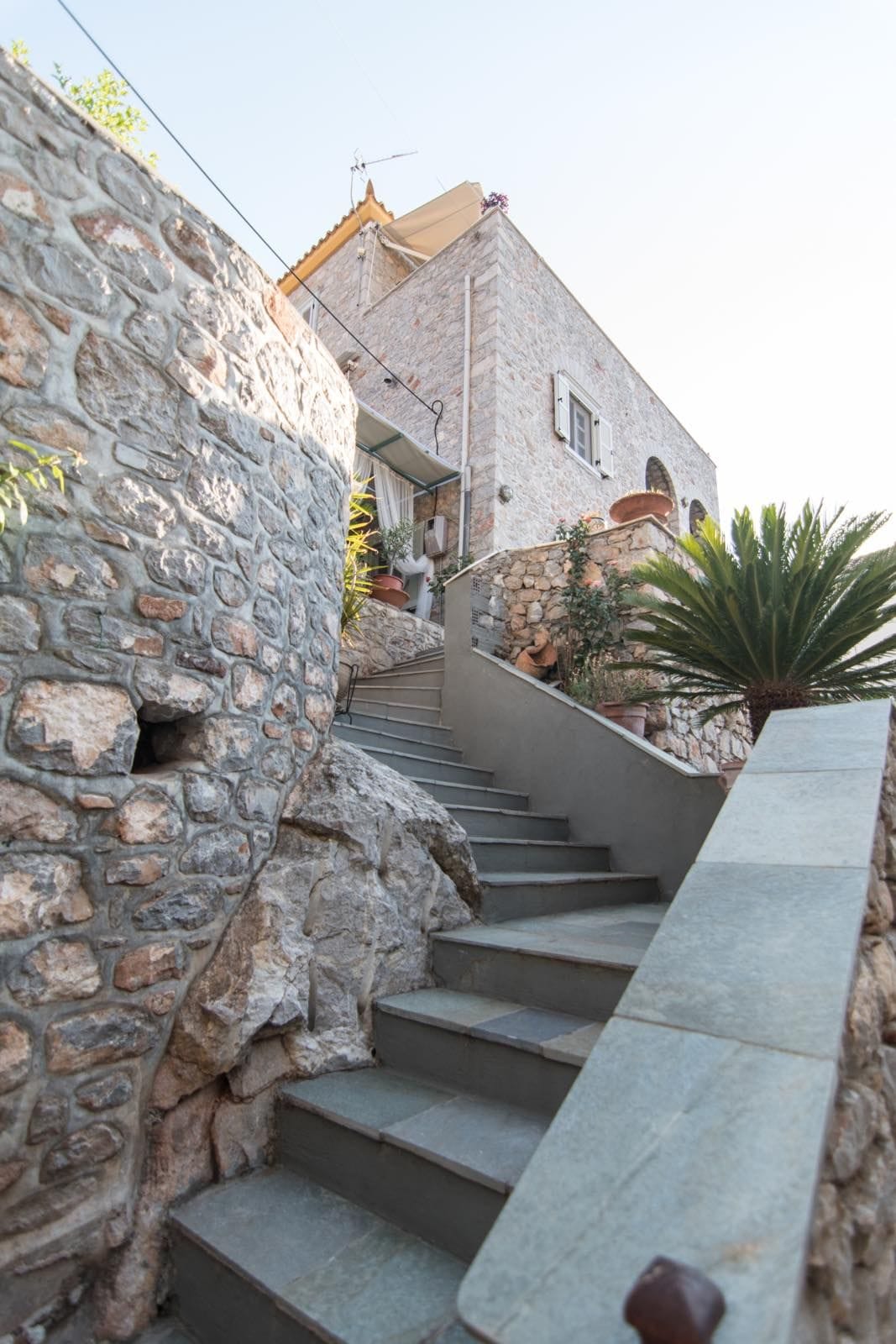 A stone staircase leads upward, flanked by a textured rock wall and lush greenery. The building's upper levels are visible, featuring a balcony with light-colored shutters. Natural light illuminates the scene, enhancing the warm tones of the stonework.