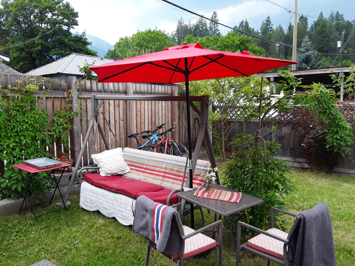 An inviting outdoor seating area features a swing lounge seat covered with a cozy blanket, accompanied by a small table and chairs. A large red umbrella provides shade, while greenery surrounds the space, creating a private and relaxing environment.