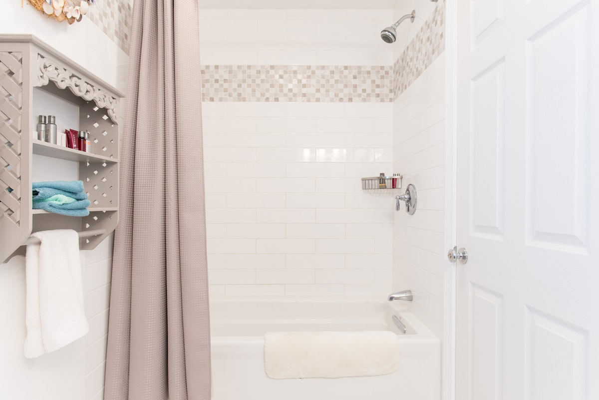 A clean and modern bathroom features a tub with a shower head overhead, surrounded by white tiled walls. A light-colored shower curtain hangs beside decorative shelves holding bath essentials and soft towels. Natural light highlights the neutral tones of the space.