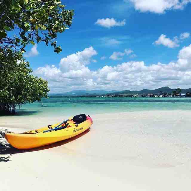 A vibrant yellow kayak rests on the sandy shore, partially surrounded by clear turquoise waters. Lush green vegetation lines the beach, and a distant horizon features rolling hills under a bright blue sky with fluffy white clouds.