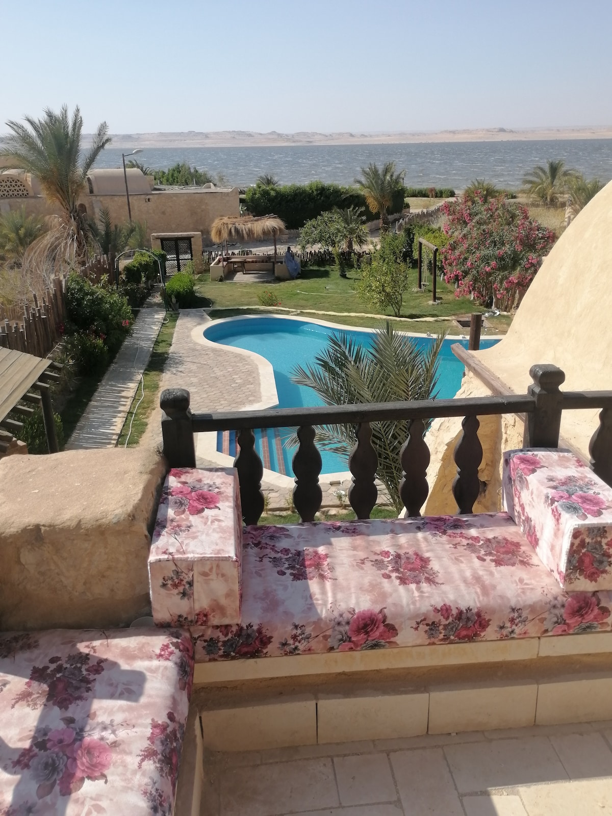 A view of a tranquil outdoor area is captured, featuring a swimming pool surrounded by lush gardens. Comfortable seating with floral upholstery is positioned on a balcony. The scene includes a serene lake and distant mountains, enhancing the natural beauty of the surroundings.