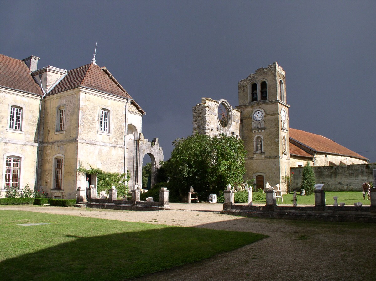A historic abbey courtyard showcases a blend of the original stone architecture and a clock tower. The grassy area is framed by weathered walls, with remnants of a church visible to one side. A sense of tranquility is present amid the surrounding greenery.