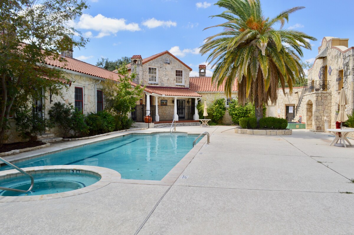 An outdoor area features a clear blue pool surrounded by laid-back seating. Palm trees and neatly trimmed shrubs add greenery to the stone architecture. A small lounge area is present beside the pool, enhancing the space for relaxation.