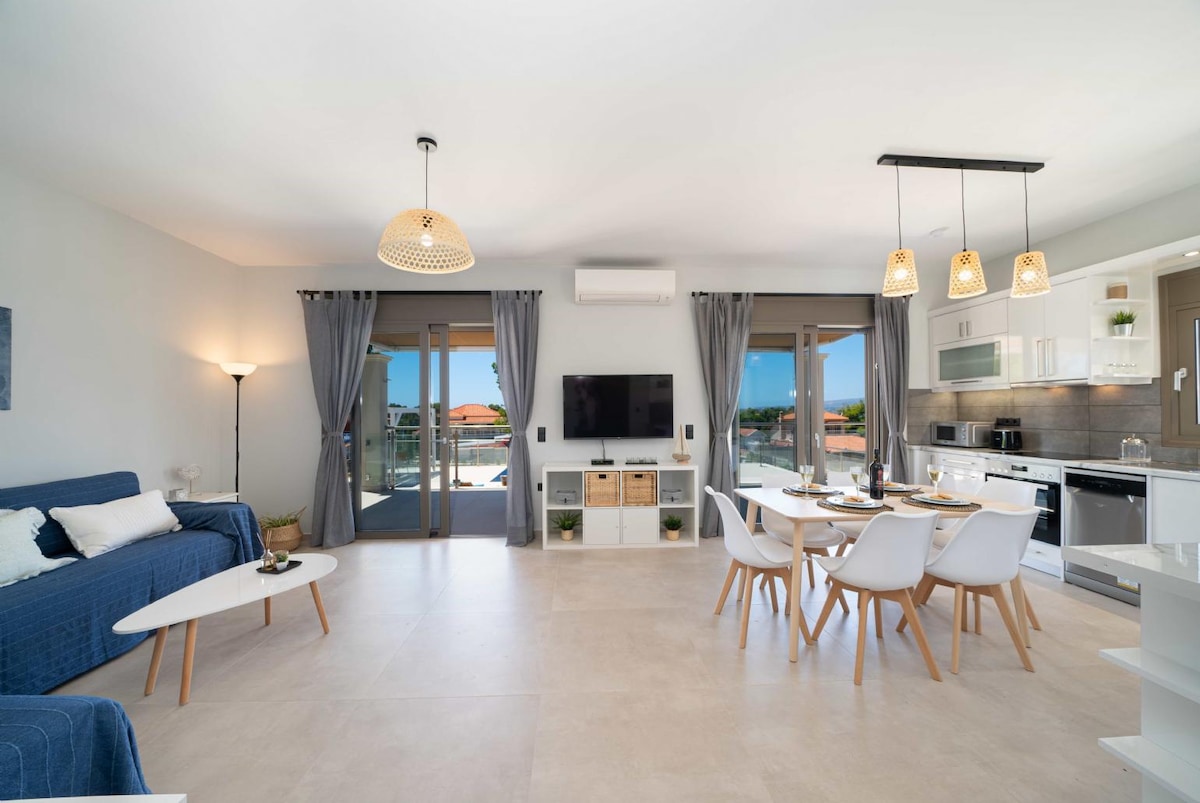 An open-plan living area is showcased, including a comfortable blue sofa and a dining table with white chairs. Large glass doors provide natural light and connect to the outdoor space. Modern fixtures and a neutral color palette create a clean and inviting atmosphere.