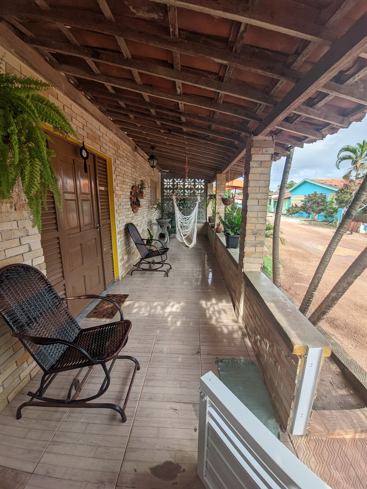A shaded porch is lined with two wicker chairs, providing a comfortable seating area. A hammock is gently displayed between the posts, enhancing relaxation. Green plants are hanging near the entrance, while the exterior showcases a rustic brick facade.