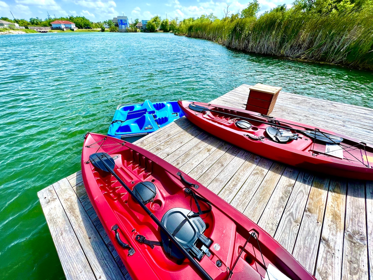 Two red kayaks, equipped with black seats, rest on a wooden dock. A blue paddle boat is visible nearby, with a serene body of water and lush greenery in the background. The scene is brightened by a clear sky and soft clouds.