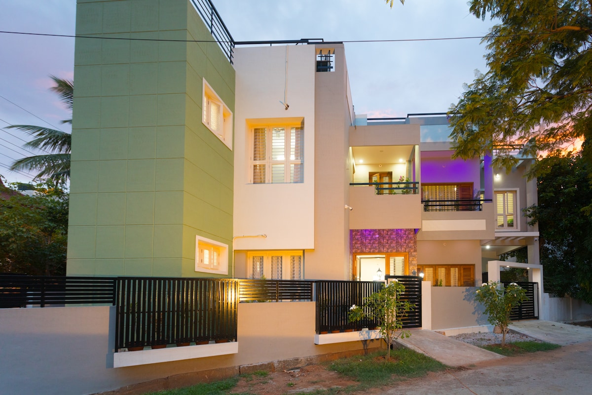 A modern guesthouse is depicted, showcasing a multi-level facade with a combination of green and neutral-colored walls. Soft lighting illuminates the windows, creating a welcoming ambiance. Lush greenery is visible along the pathway leading up to the entrance, complementing the residential setting.