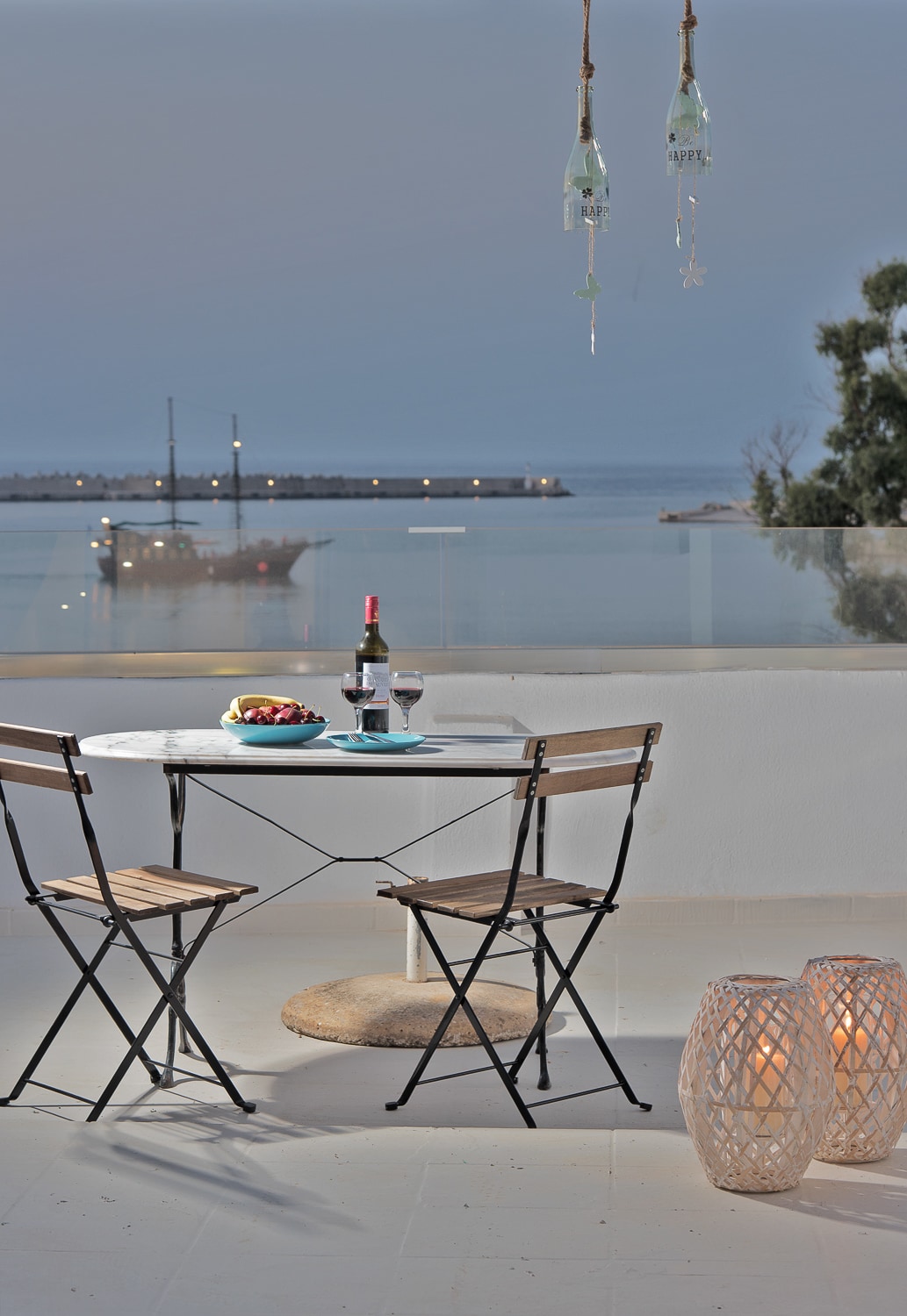 A dining table for two is set on a balcony overlooking the sea. Two folding chairs frame the table, which features a bottle of wine and glasses. Softly lit lanterns add warmth to the scene, while a view of the harbor is visible in the background.