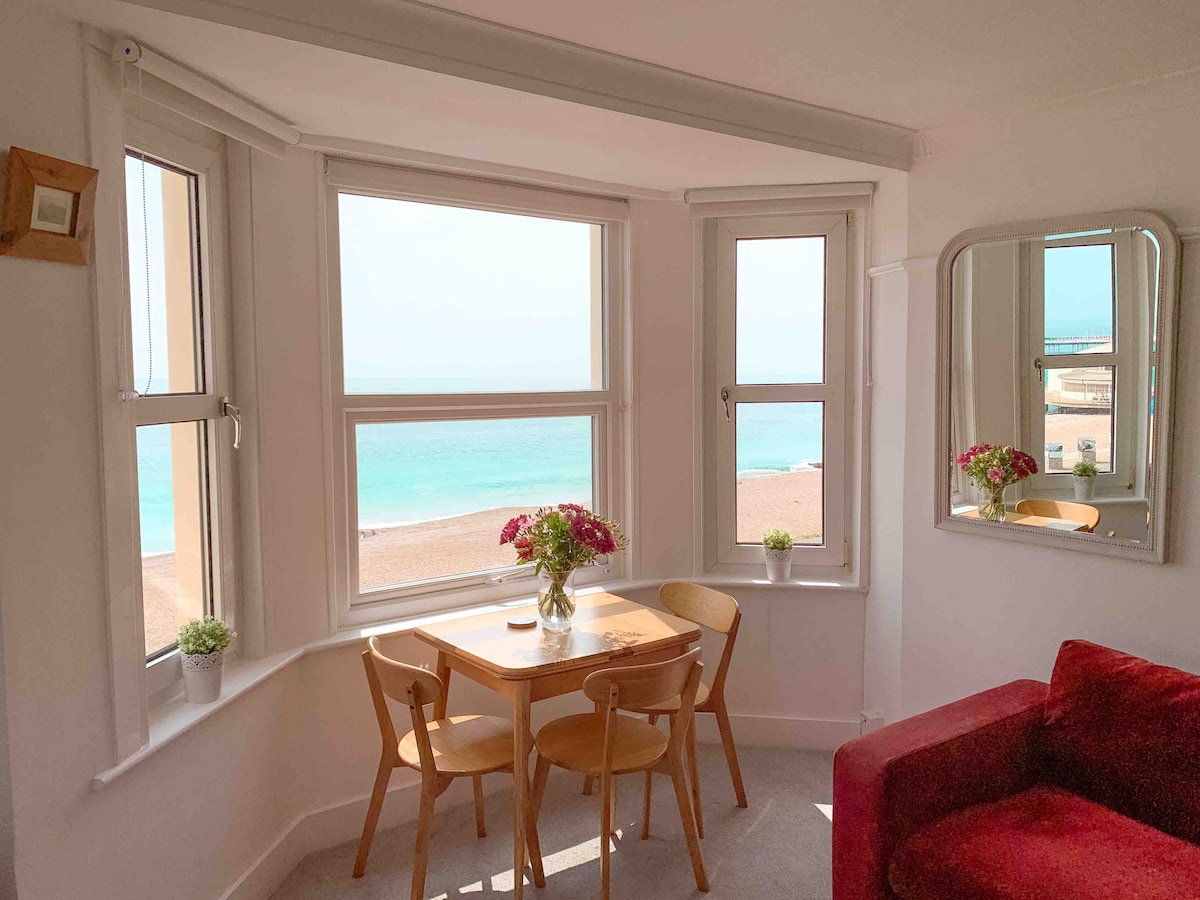 A cozy dining area features a small wooden table surrounded by four chairs, positioned by large windows with sea views. A vase with flowers rests on the table, while natural light brightens the space. A mirror reflects the inviting atmosphere.