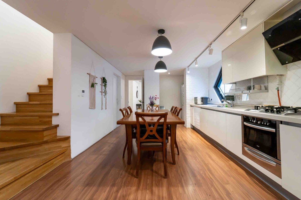 A modern open-concept kitchen and dining area is showcased, featuring a wooden dining table surrounded by six chairs. Natural light enters through a unique diamond-shaped window. Warm wooden floors complement the sleek cabinetry and appliances, while a staircase leads to an upper level.