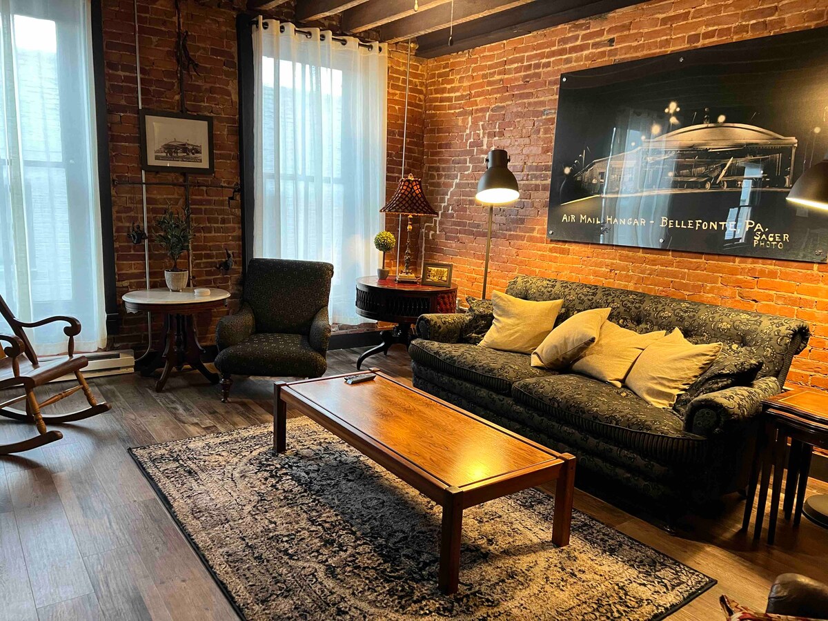 A cozy living area features exposed brick walls and wooden beams, with natural light illuminating the space. A comfortable sofa with decorative pillows faces a wooden coffee table, while a rocking chair and a small table provide additional seating options.