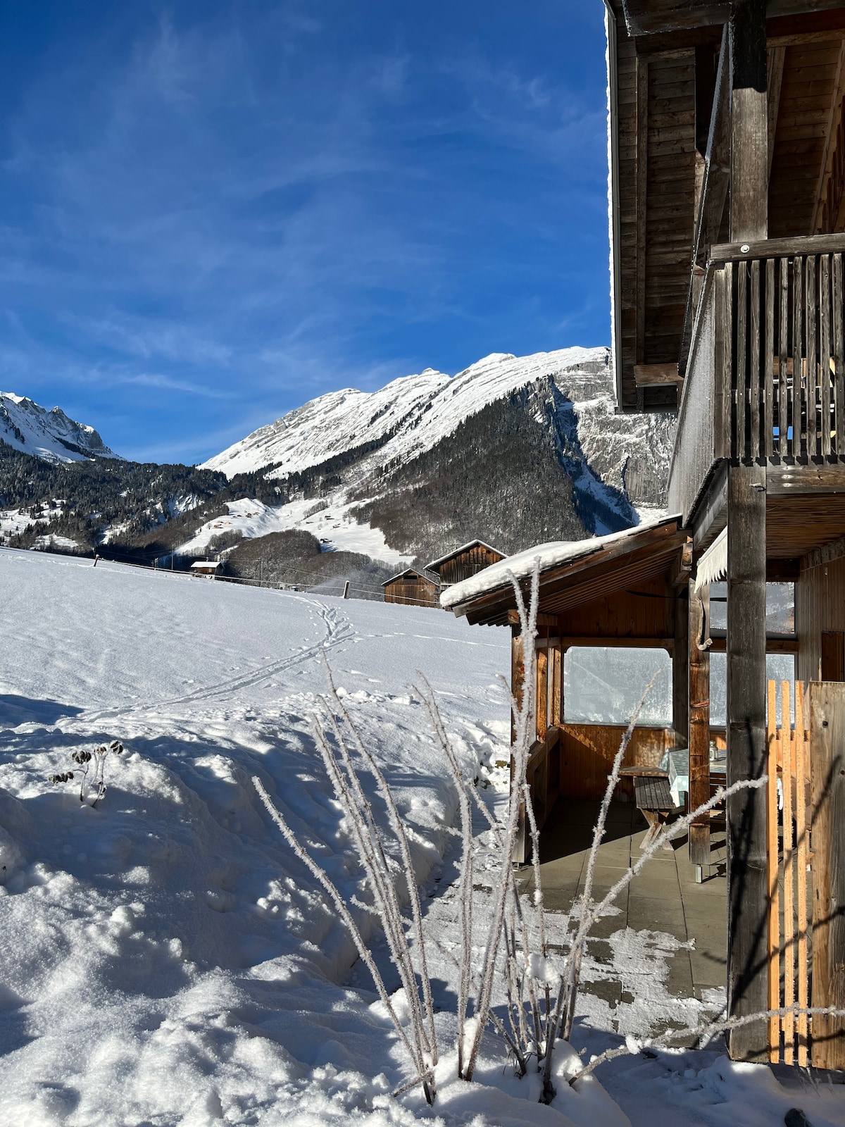 A snow-covered landscape is seen alongside a traditional wooden house. The surrounding mountains rise majestically in the background, under a clear blue sky. A pathway through the snow leads towards the horizon, offering a sense of tranquility and winter beauty.