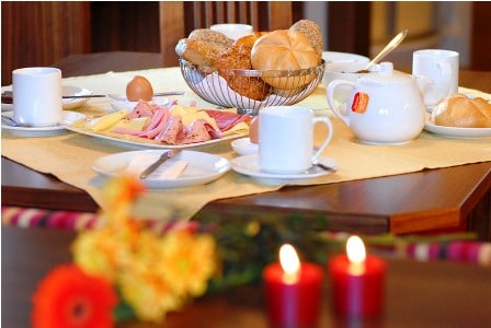 A dining table is set with a variety of breakfast items, including a woven basket of bread rolls and a selection of sliced meats and cheeses. Two cups and a teapot are positioned alongside a plate with an egg. Fresh flowers and lit candles add subtle touches.