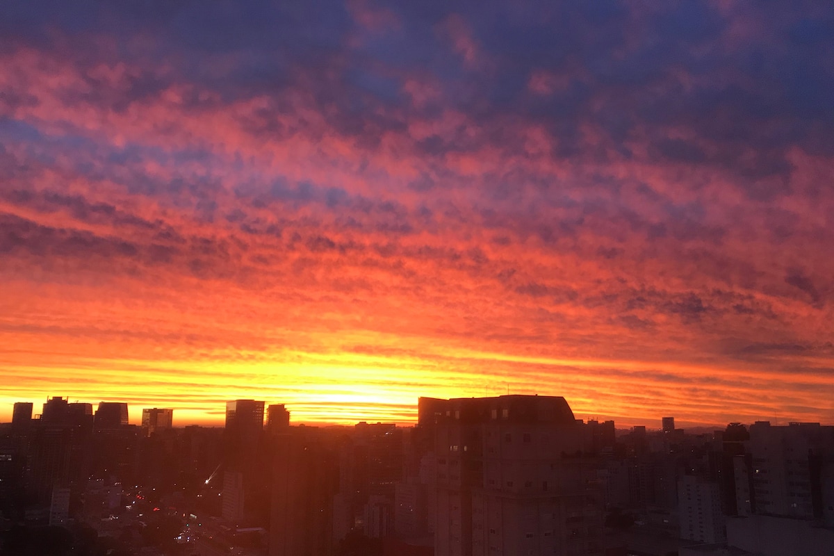 A vibrant sunset paints the sky in shades of orange, pink, and purple over the São Paulo skyline. Buildings silhouette against the colorful horizon, creating a serene urban scene. This captivating view offers a glimpse of the city's dynamic landscape transitioning from day to night.