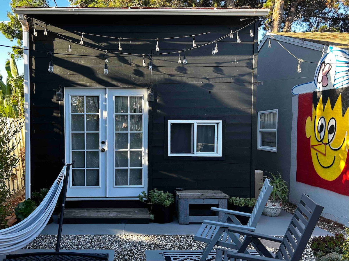 The exterior of a charming black cottage is displayed, featuring double French doors framed by large windows. A cozy seating area with wooden chairs is present on a patio adorned with gravel, while string lights hang above, adding a welcoming touch.