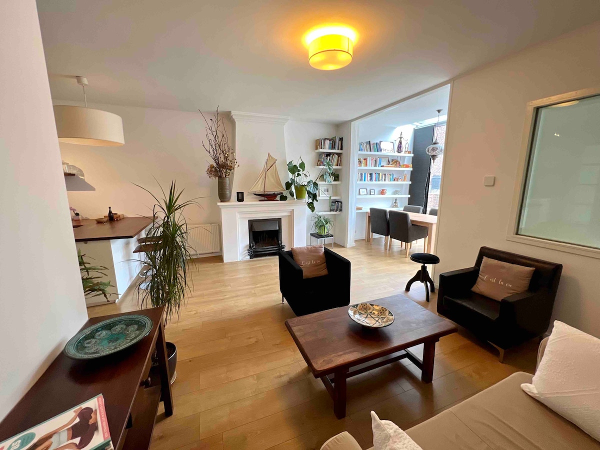 An airy living space is displayed, featuring wooden flooring and a fireplace. A coffee table sits in the center, surrounded by a sofa and armchair. A small dining area is visible, adorned with plants. Bookshelves and a decorative sailboat add character to the room.
