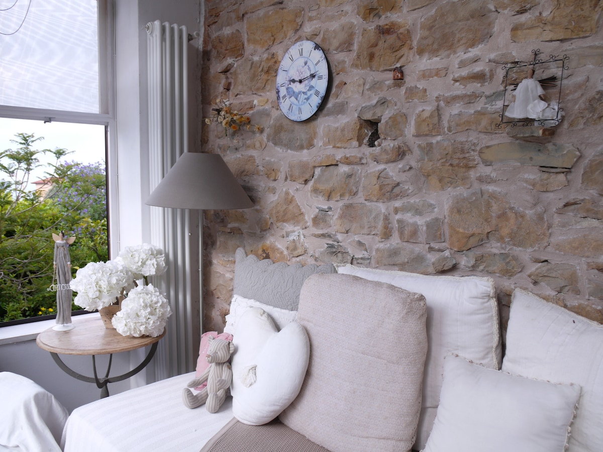 A cozy corner showcases a stone wall, complemented by a large window allowing natural light to illuminate the space. A soft beige sofa is adorned with various cushions and a decorative stuffed animal. A round table holds a vase of white flowers, enhancing the inviting environment.