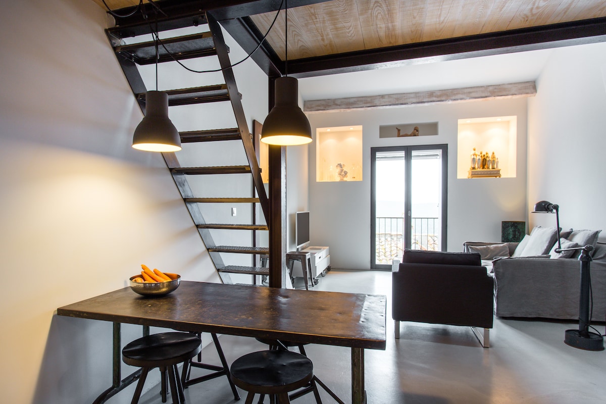 A modern open space features a rustic dining table with dark wood and three black stools, inviting communal meals. Above, two pendant lights hang from the ceiling. A staircase leads to the mezzanine. A comfortable seating area lies adjacent, with large windows providing natural light.